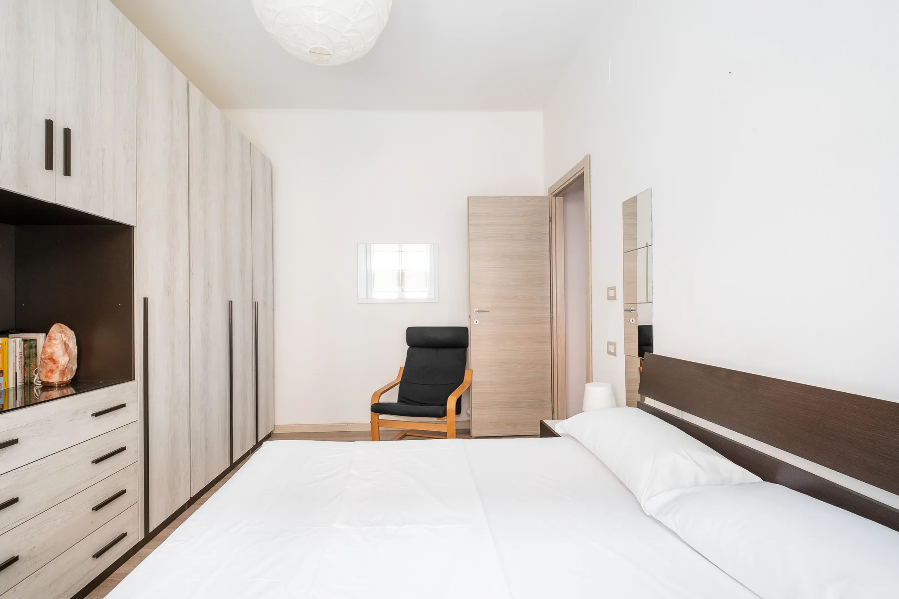 A bright bedroom with a neatly made bed, built-in wardrobes, and a chair.