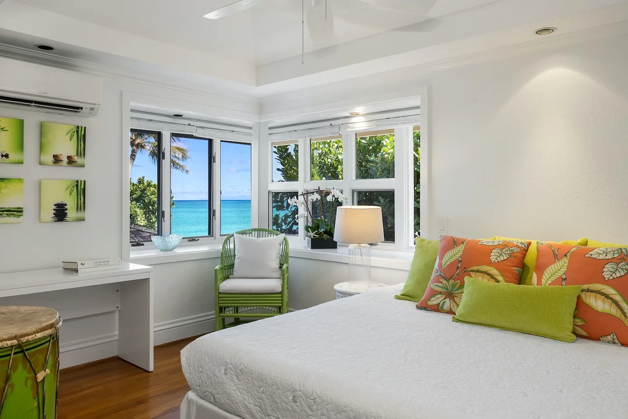 A bright bedroom with a white bed beside large windows overlooking the ocean.