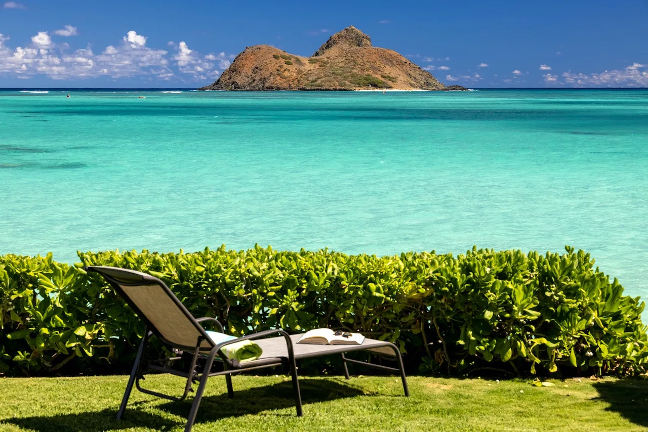 A lounge chair sits on a grassy lawn beside turquoise water and a distant island.