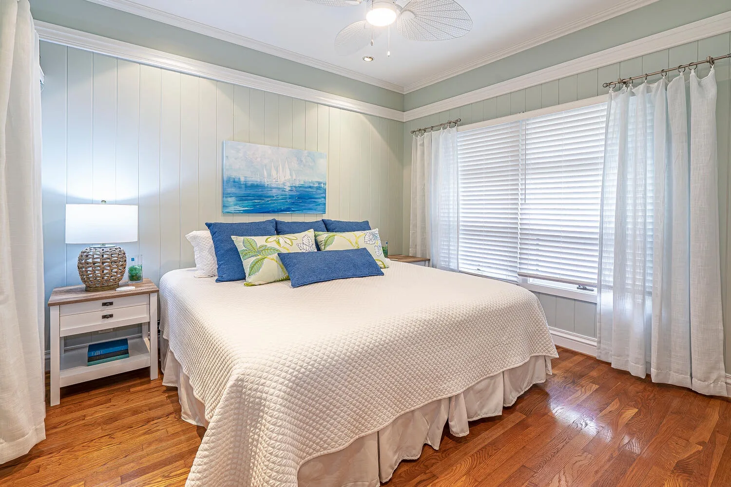 A bright bedroom with a large bed and wide windows covered by blinds.