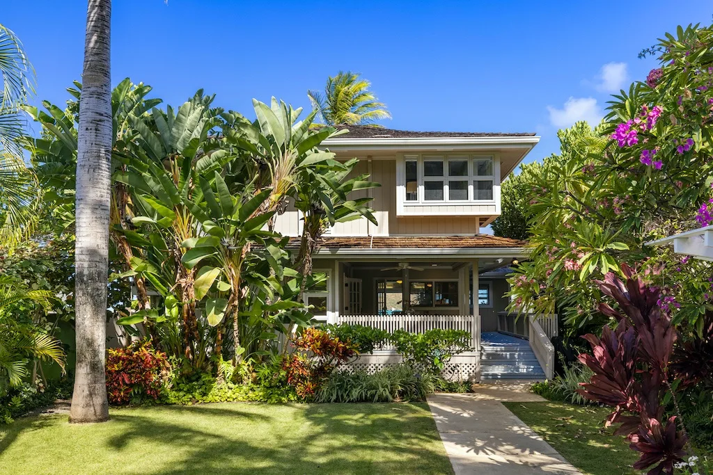 A two-story house stands behind lush tropical landscaping and a paved walkway.