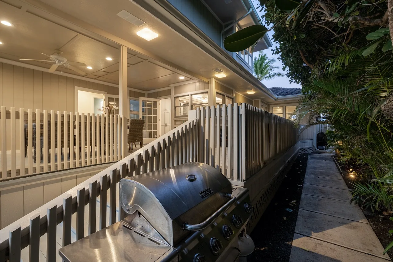 A covered outdoor patio with seating overlooks a narrow paved walkway beside a grill.