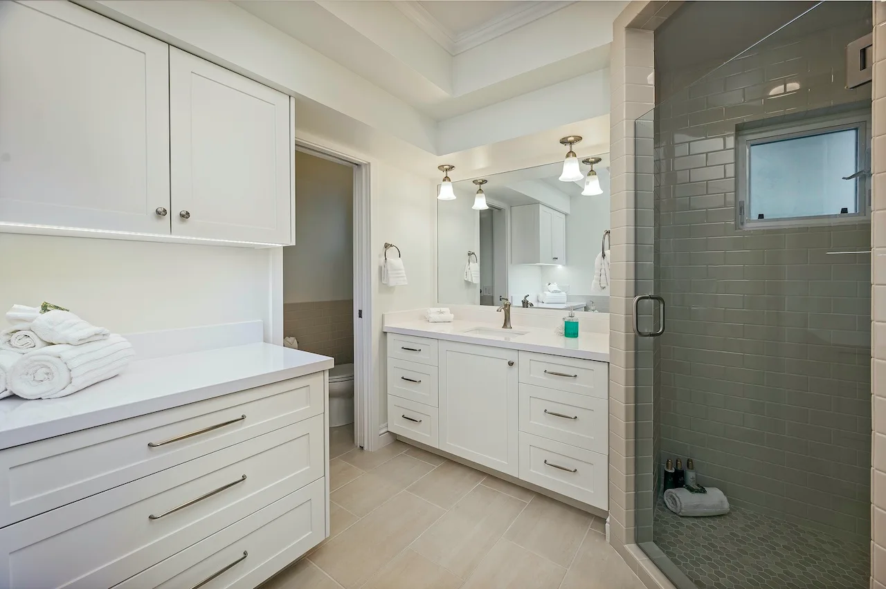 A bright bathroom with white vanities and a tiled shower.