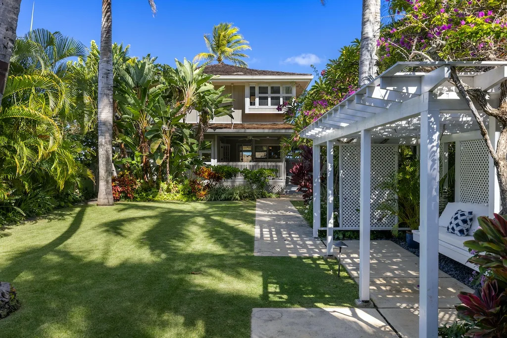 A tropical garden lawn and white pergola sit in front of a two-story house.