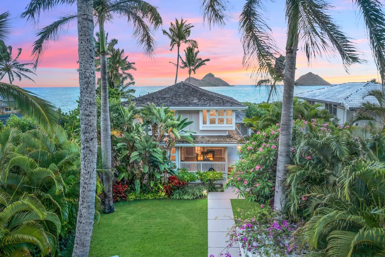 A tropical house stands among palms and flowering plants beside the ocean at sunset.