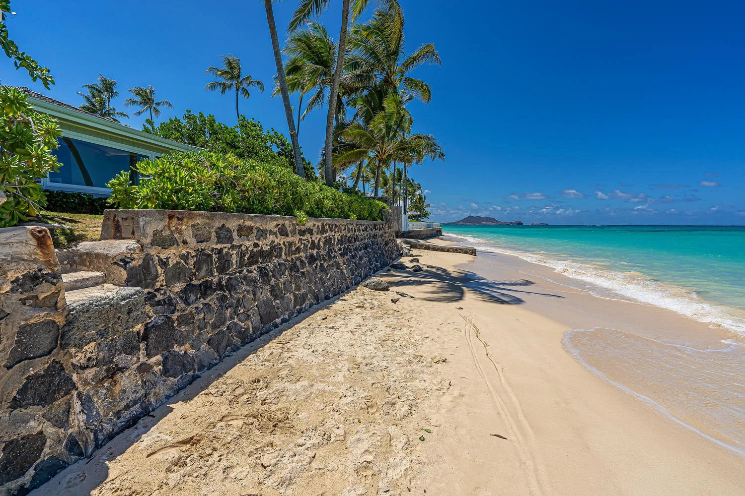 A tropical beach with pale sand, turquoise water, palm trees, and a stone wall.
