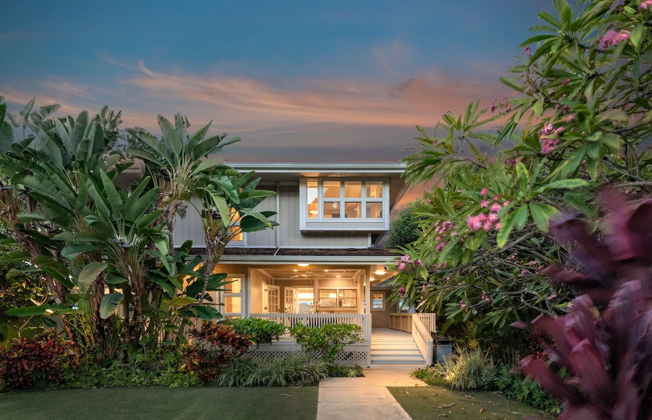 A two-story house stands behind a garden path and lush tropical plants.