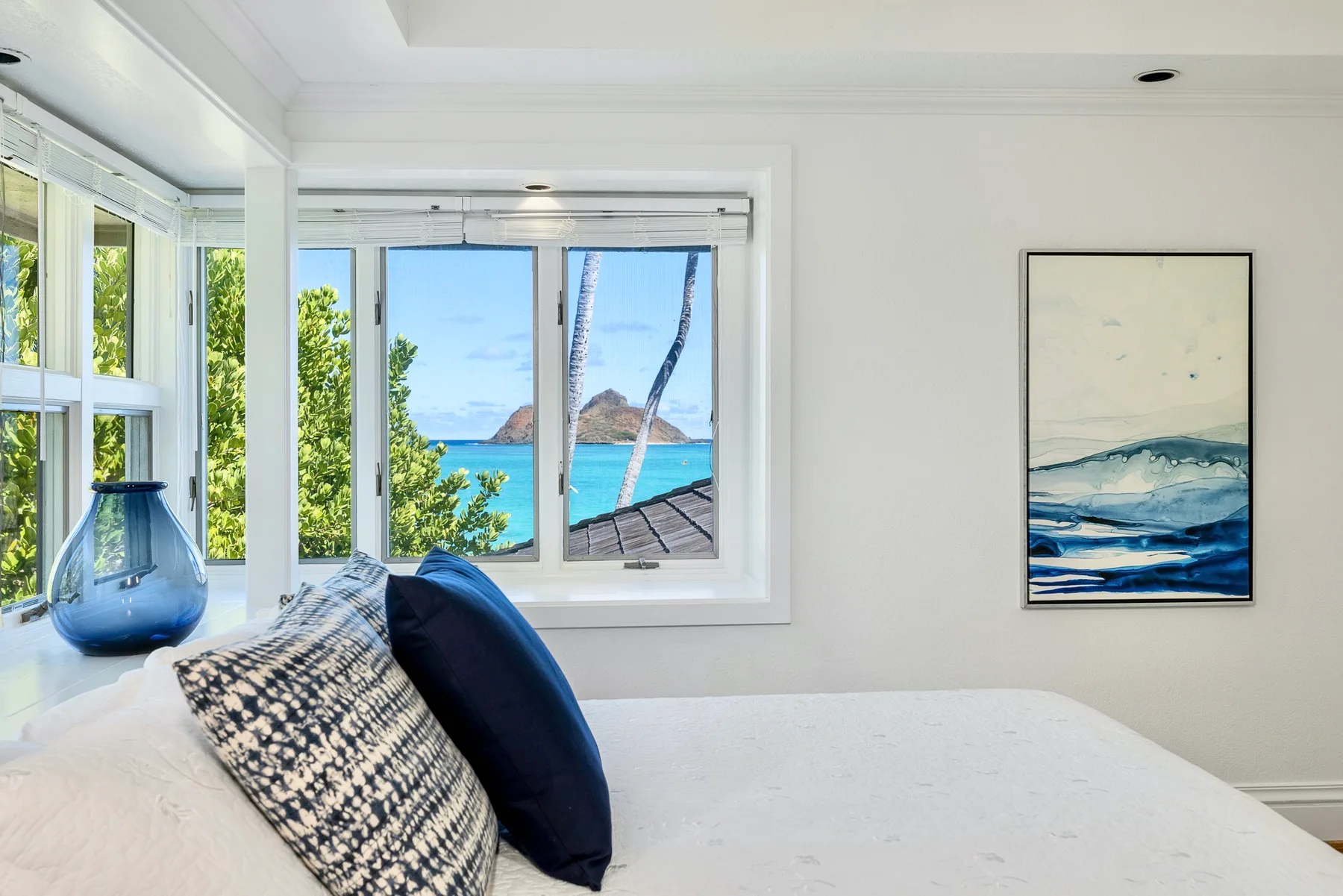 A bright bedroom with a bed beside large windows overlooking the ocean.