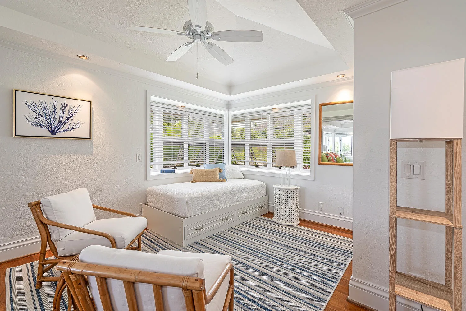 A bright bedroom with a bed beneath corner windows and a striped rug.