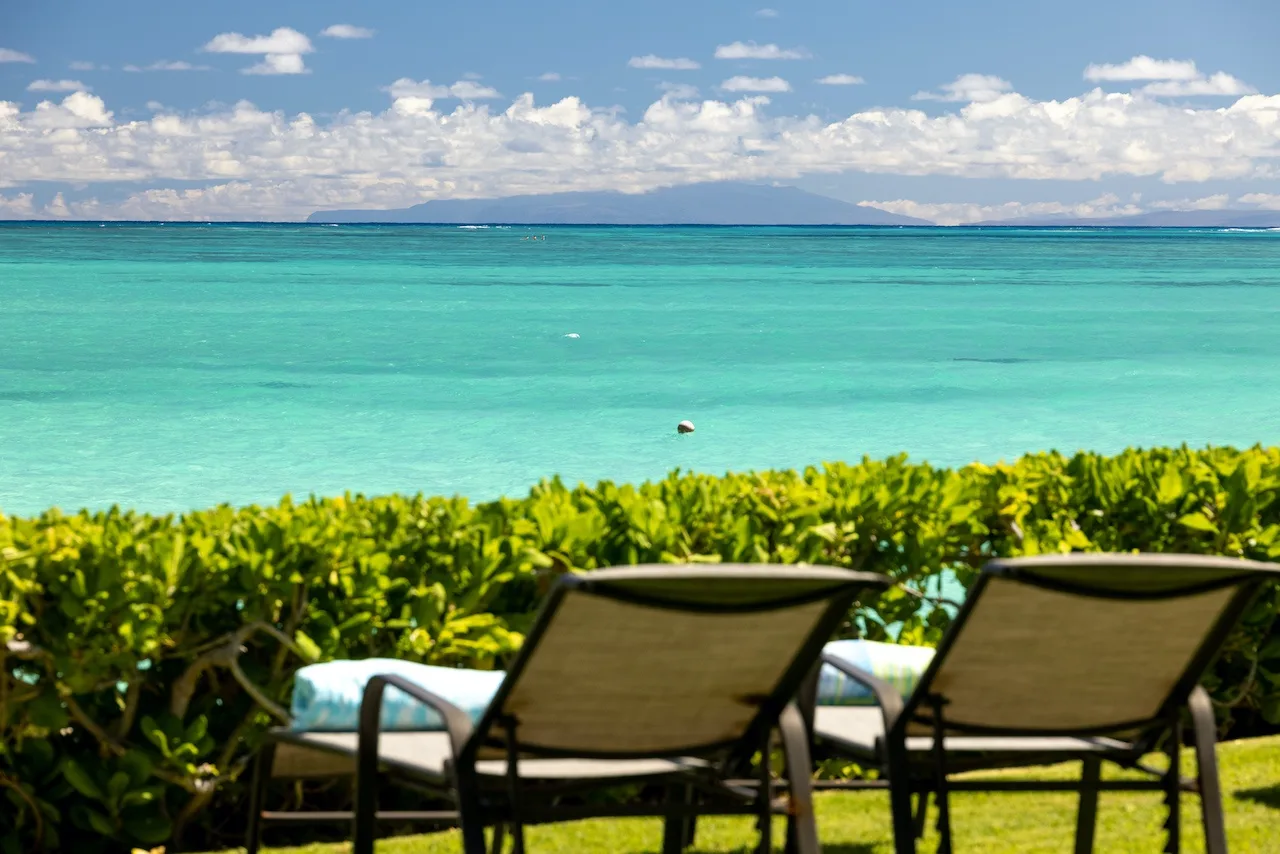 Two lounge chairs face turquoise water beyond a hedge.
