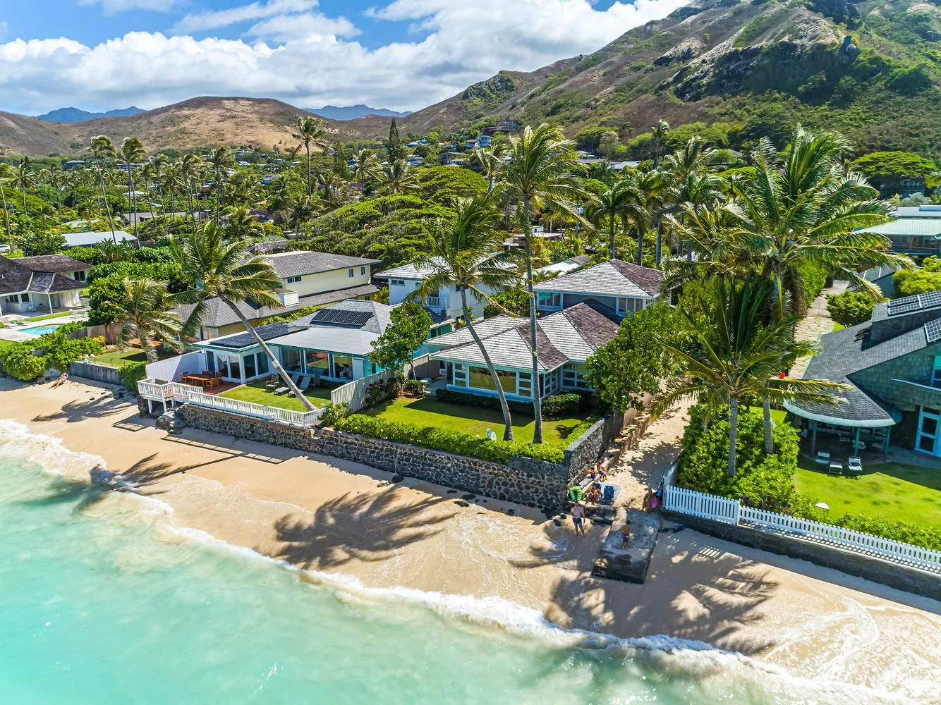 Aerial view of beachfront homes along a sandy shore, with mountains in the background.