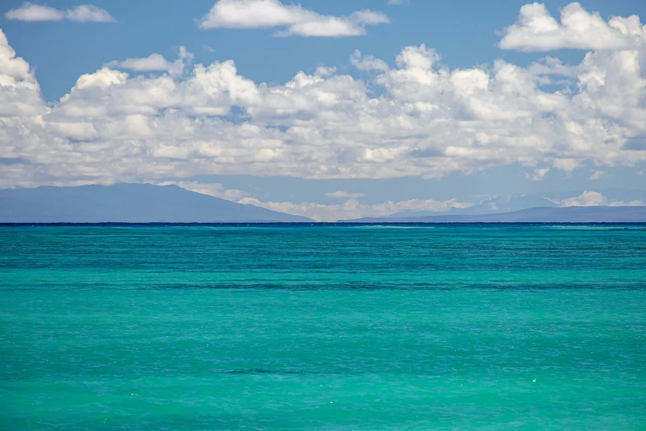 A turquoise sea stretches toward distant mountains beneath a cloudy sky.