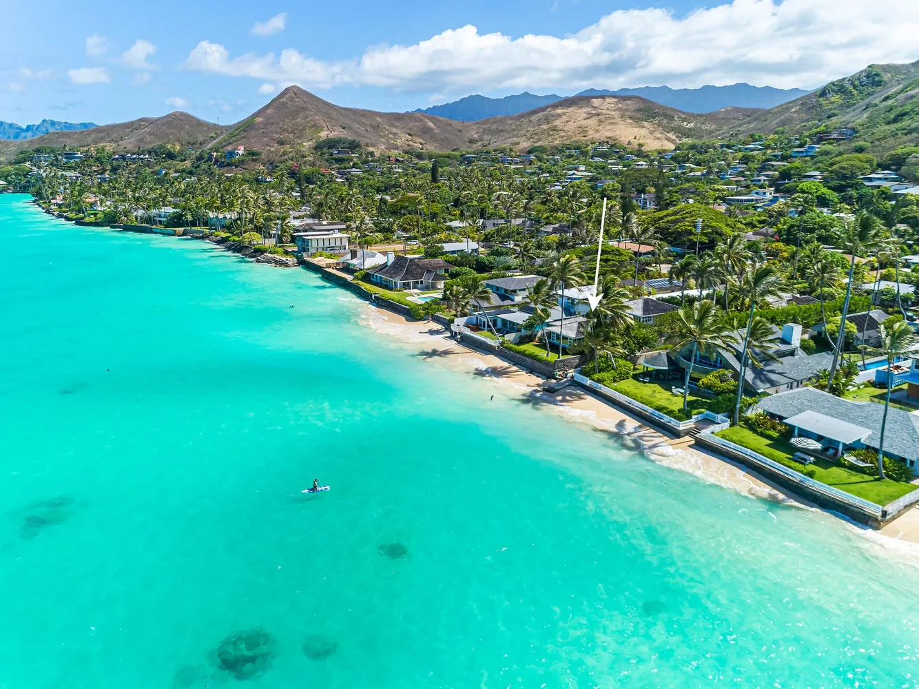 Aerial coastal scene with turquoise water, a sandy shoreline, and homes among green hills.