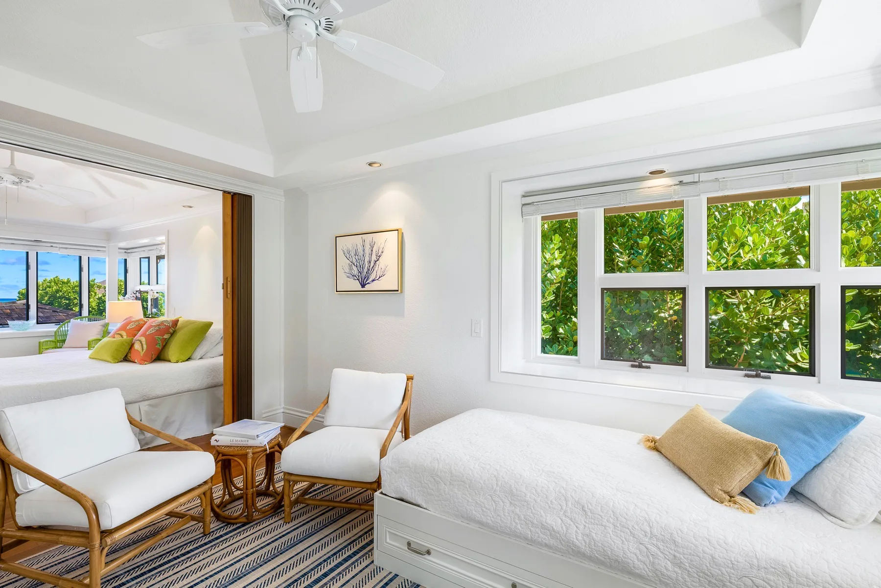 A bright bedroom with a white bed beside large windows overlooking leafy greenery.
