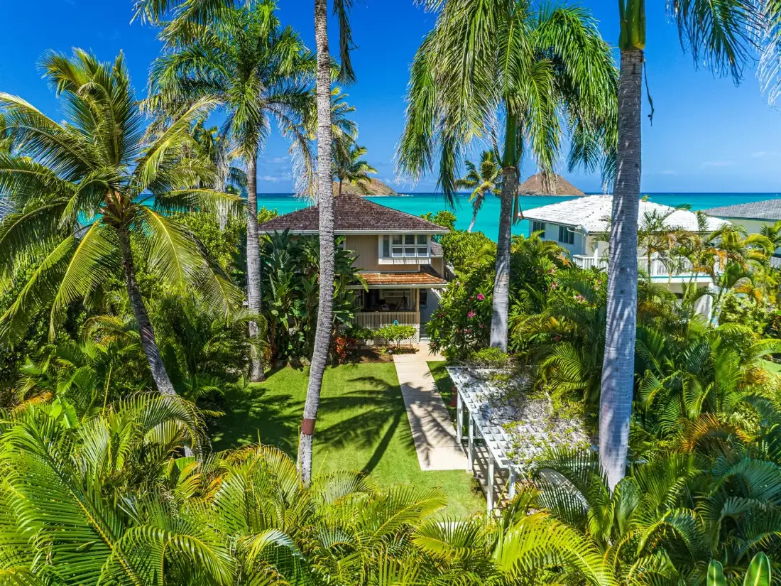 A tropical house is surrounded by palms, with the ocean visible beyond.