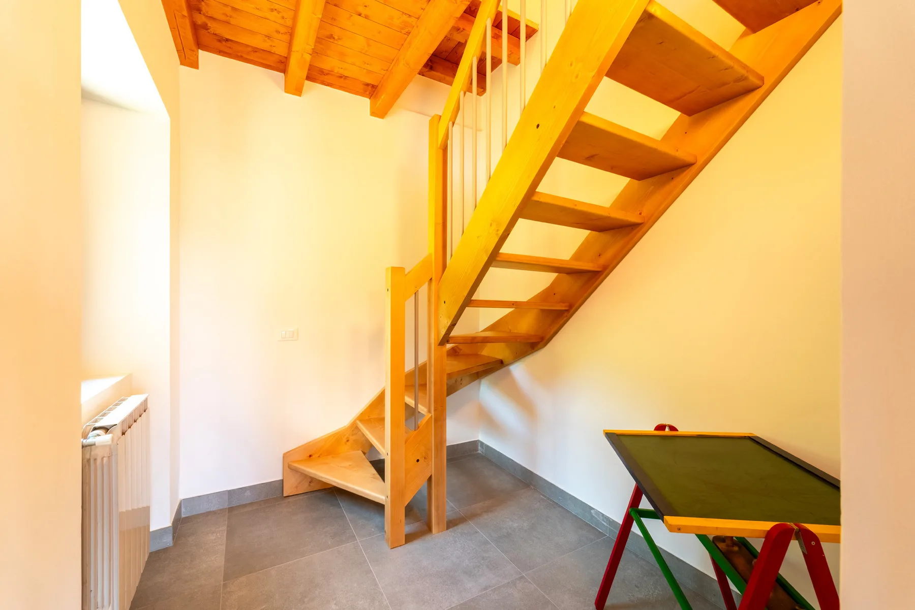 Una escalera de madera sube por un interior luminoso, con una mesa pequeña debajo.