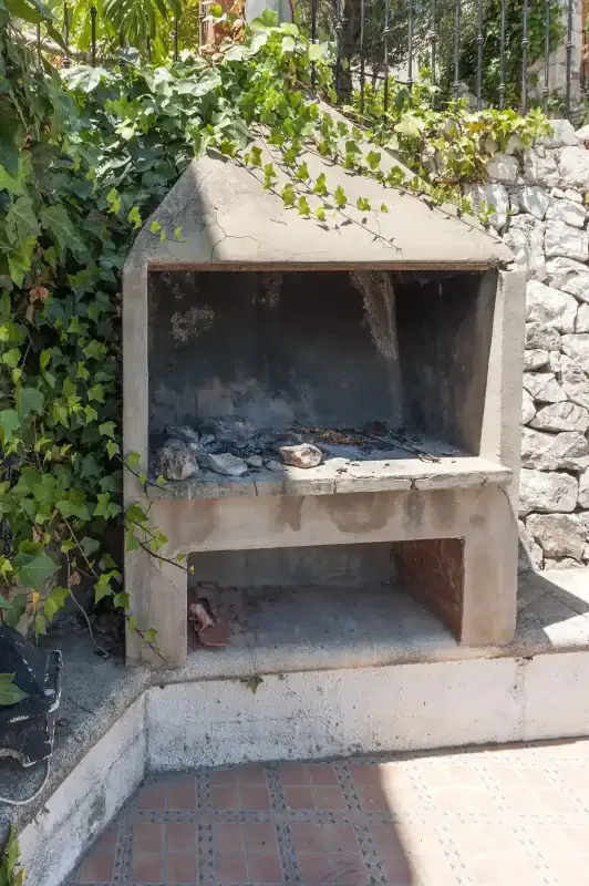 An outdoor masonry barbecue stands beside a stone wall, partly covered with vines.