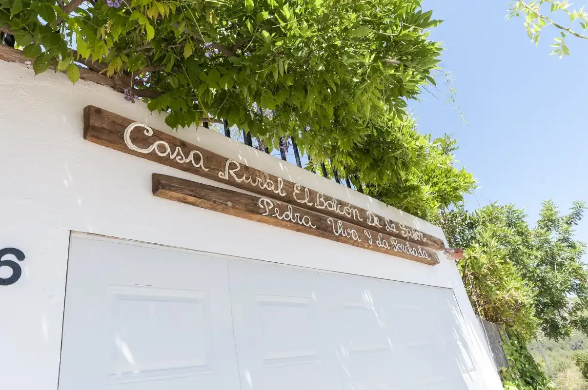 A white garage door sits beneath leafy branches, with a wooden sign above it.