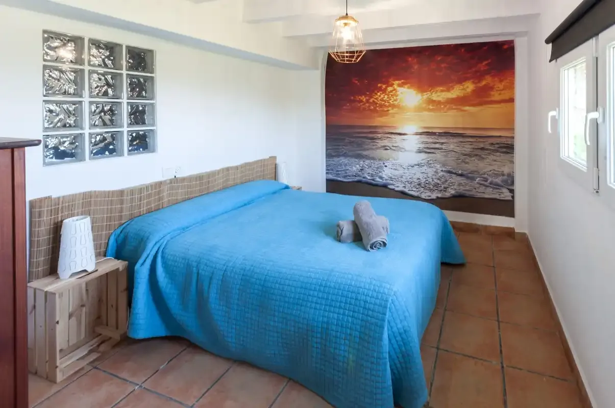 A bedroom with a blue bedspread and a large sunset beach image on the wall.