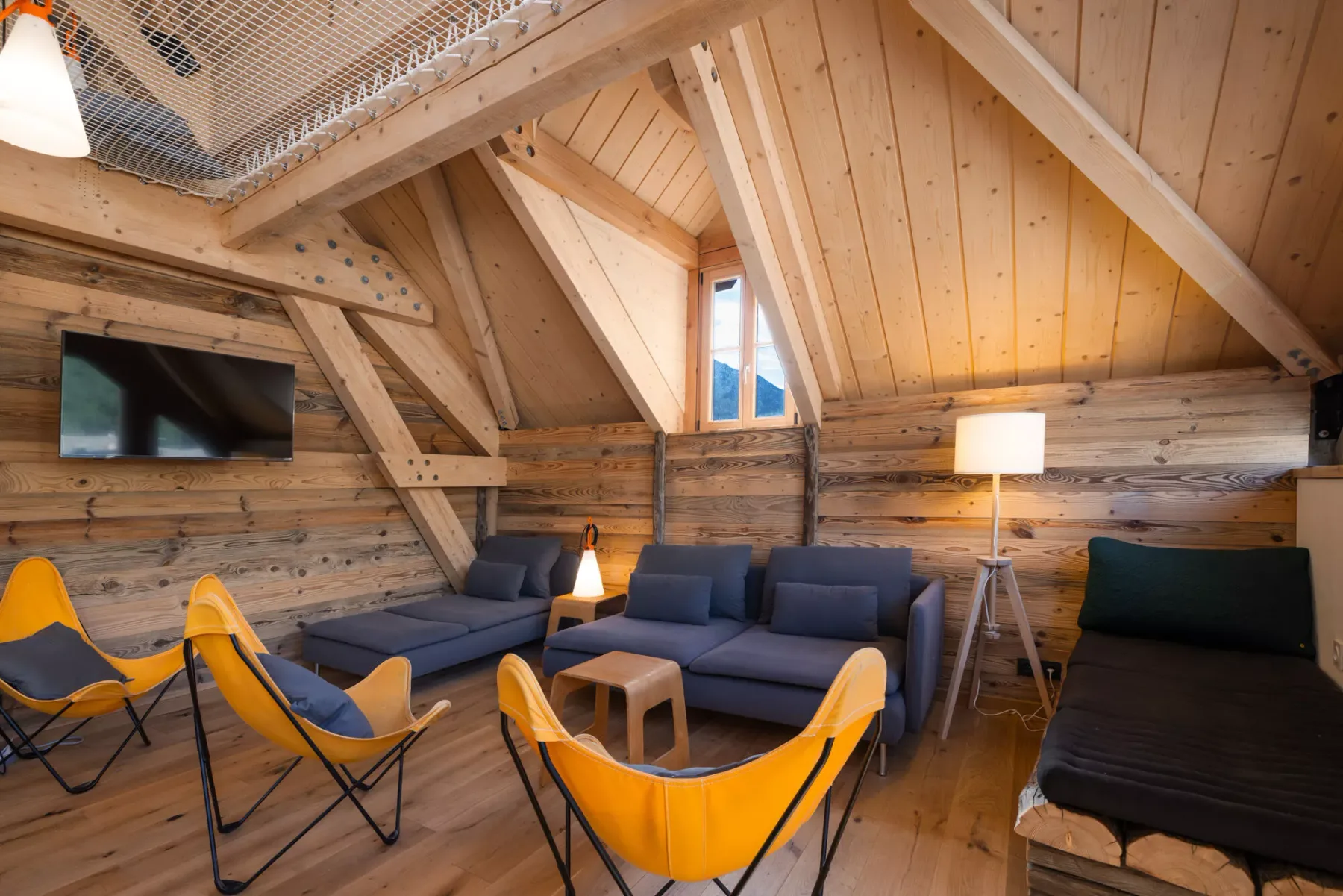 A timber loft lounge with blue seating, bright yellow chairs, and a wall-mounted television.