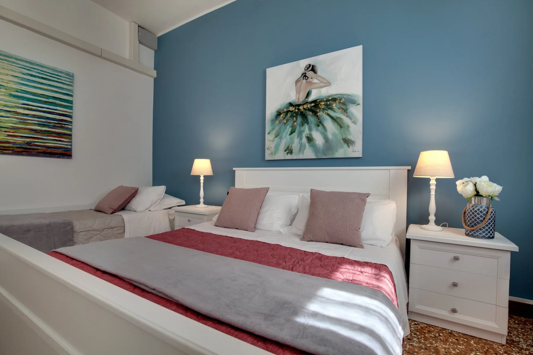 A bedroom with a large bed, white nightstands, and artwork on a blue wall.