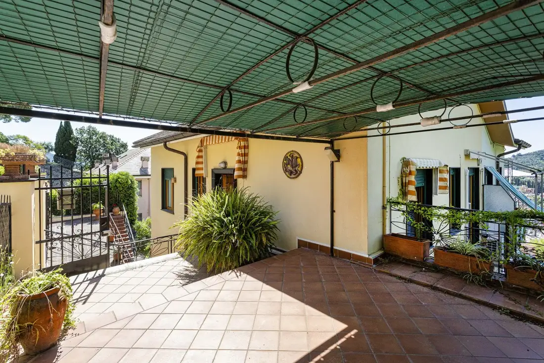 Una terraza exterior con suelo de baldosas queda bajo un toldo verde, junto a plantas en macetas y una puerta.