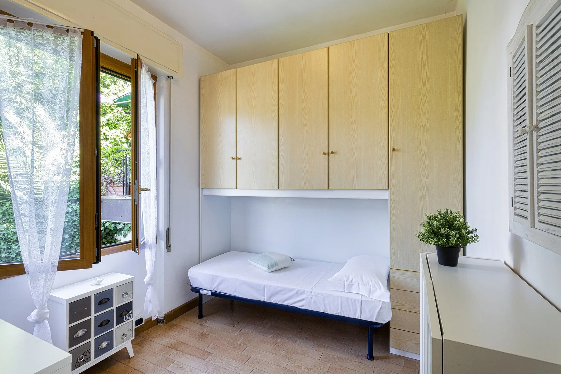 Dormitorio con una cama individual bajo armarios de madera clara y ventanas a un lado.