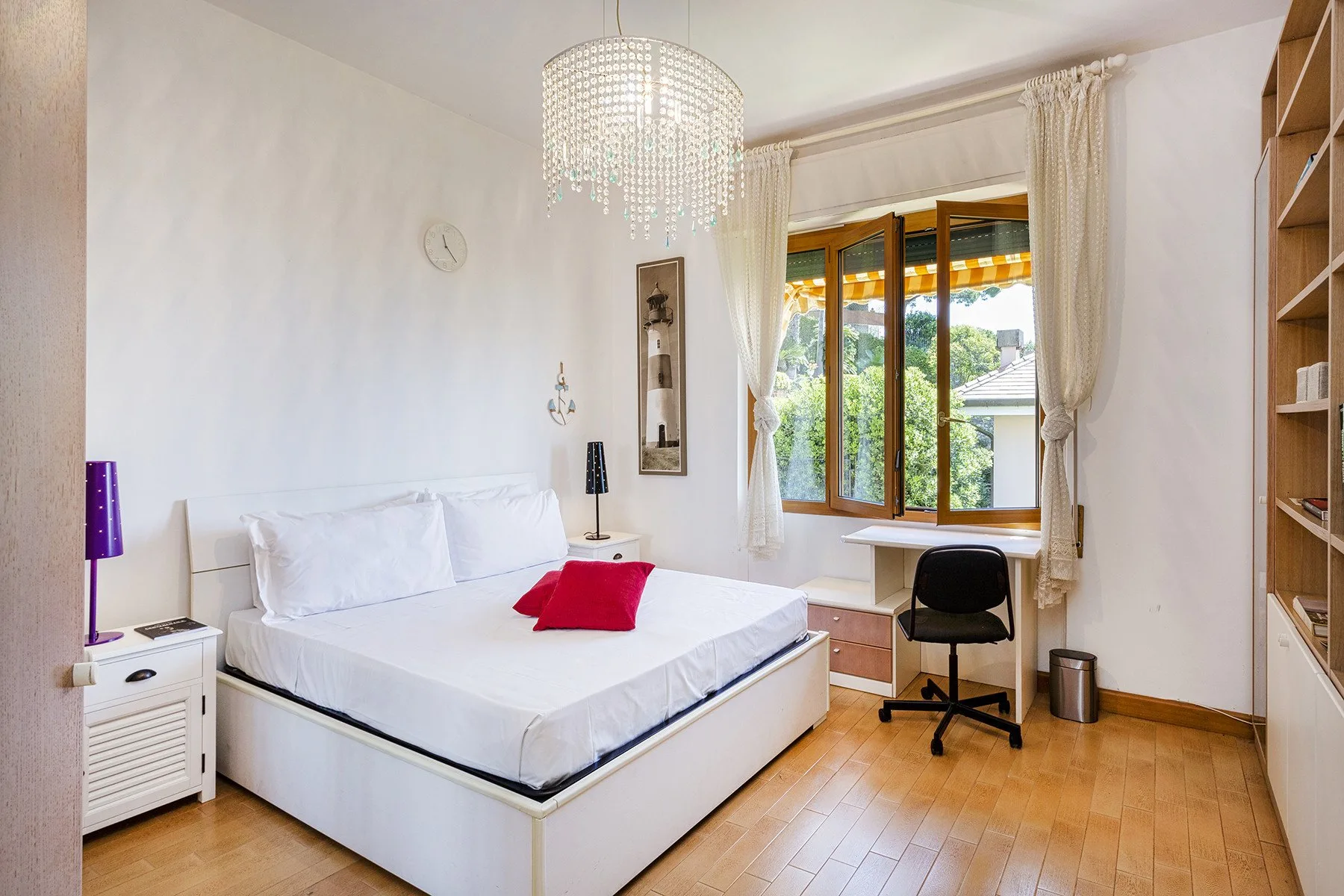 Un dormitorio luminoso con una cama blanca, mesitas de noche y un escritorio junto a la ventana.