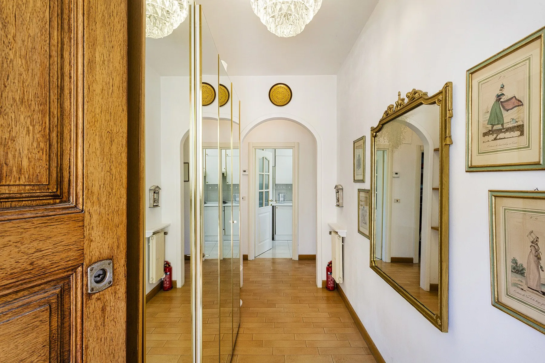 Un pasillo luminoso con un espejo grande de marco dorado y una puerta arqueada.