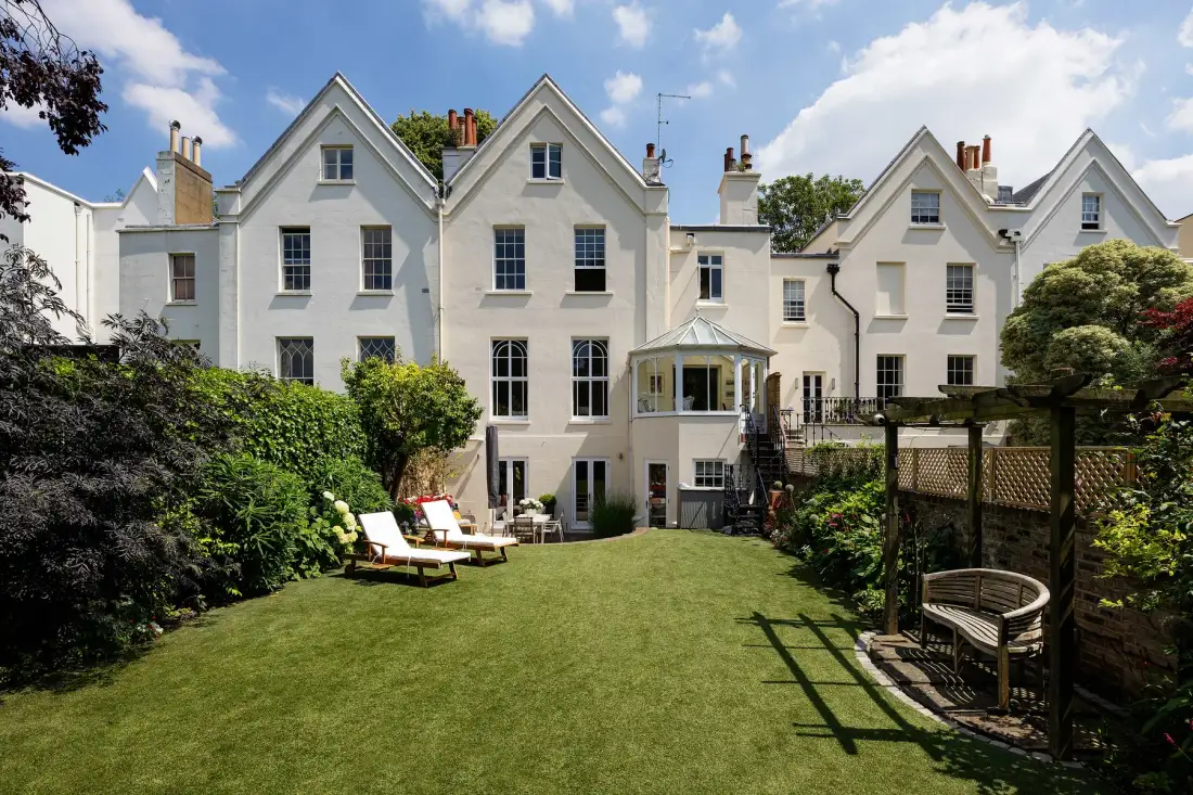 A white multi-gabled house overlooks a green garden with outdoor seating.