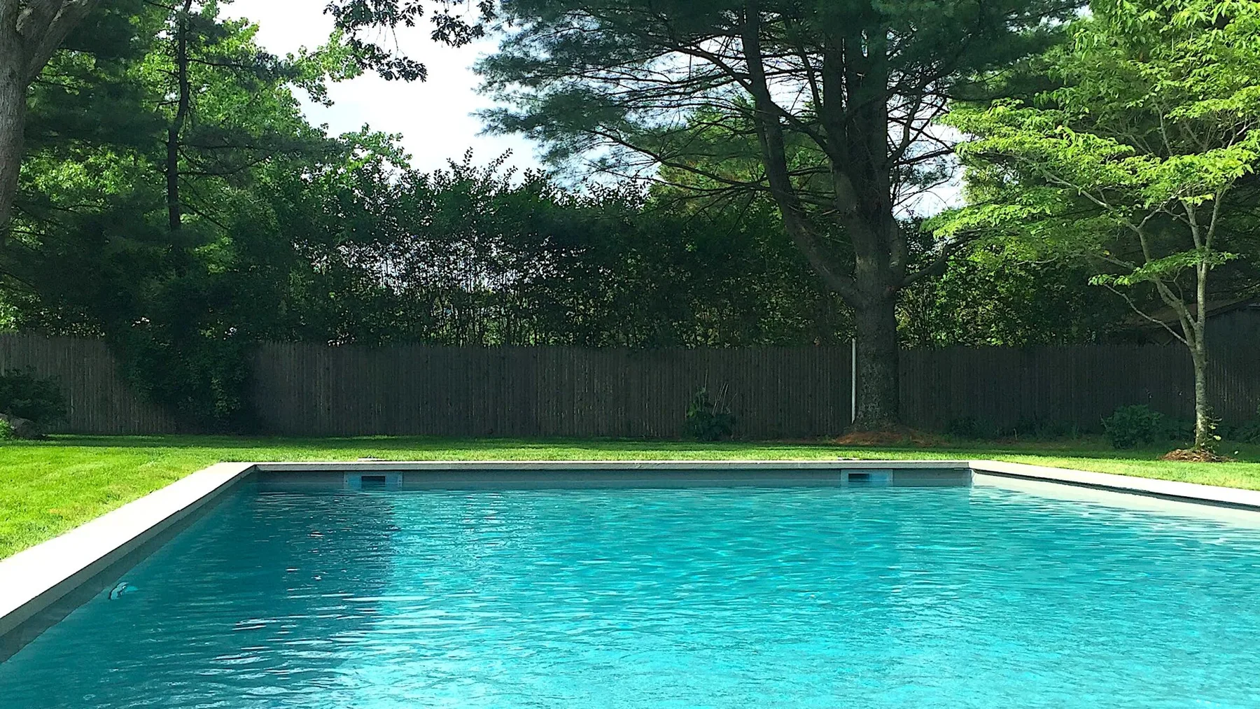 Una piscina al aire libre junto a un césped y árboles frondosos.