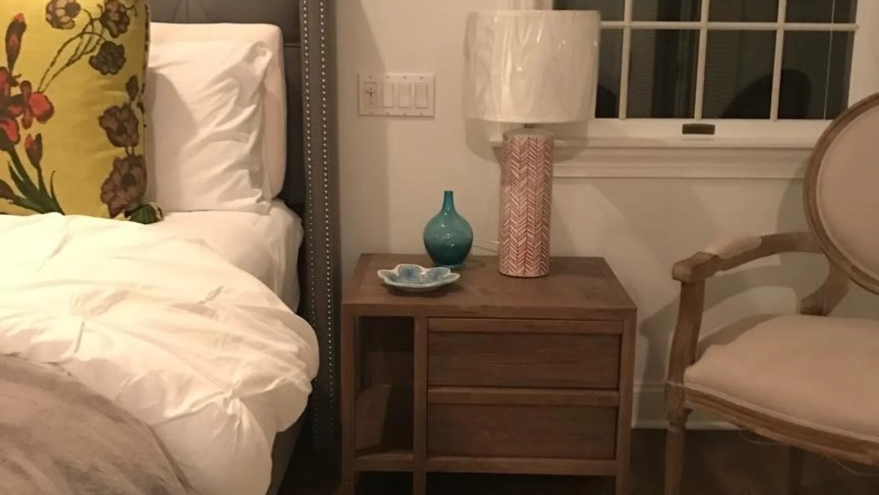 Un dormitorio con cama, mesita de noche y silla junto a una ventana.