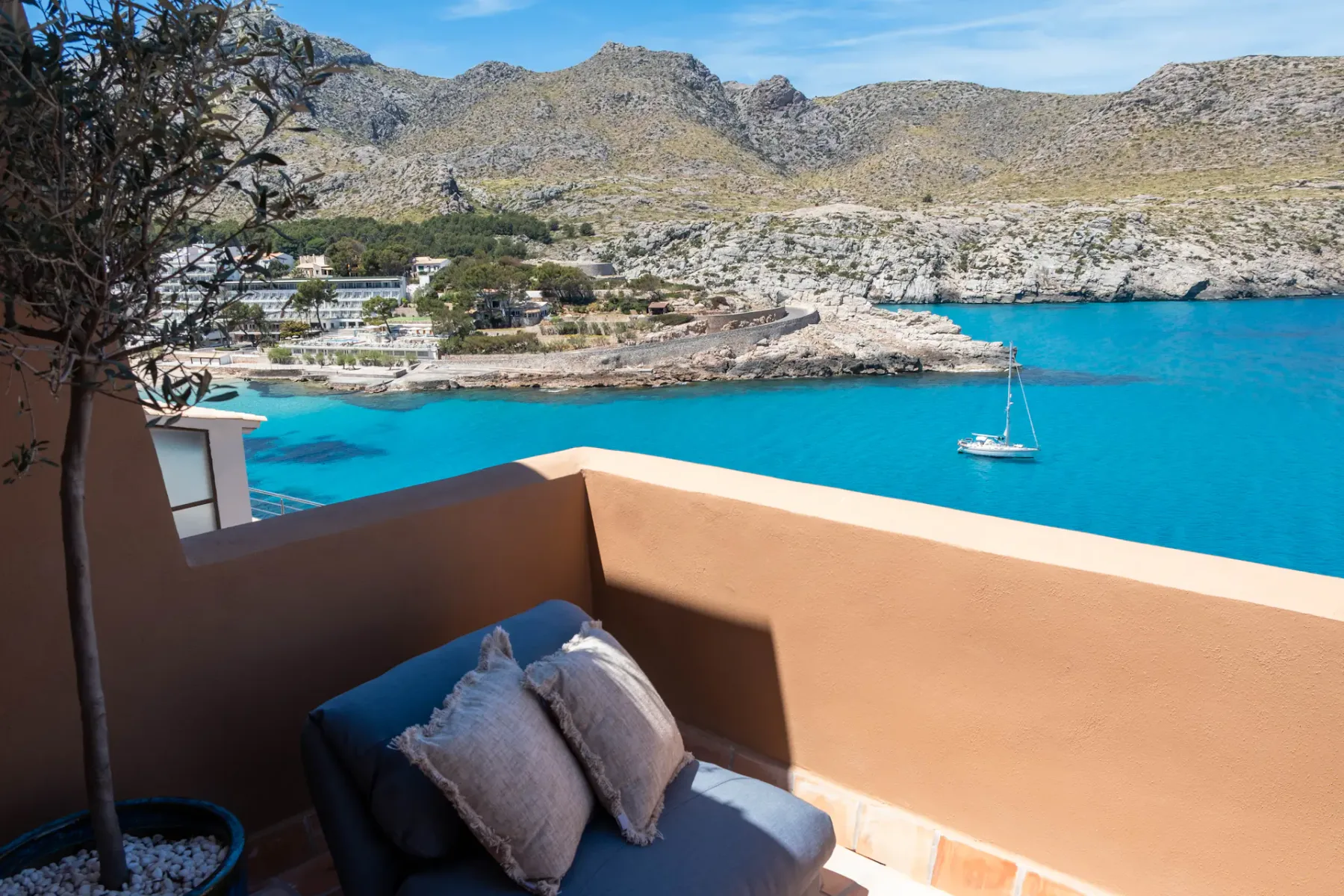 A balcony seating area overlooks a bright blue bay and rocky hills.