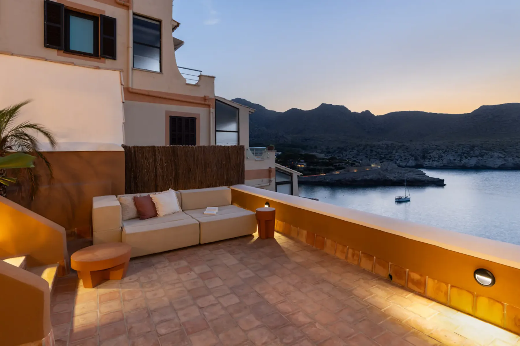 A terrace with outdoor seating overlooks a bay and surrounding mountains at dusk.