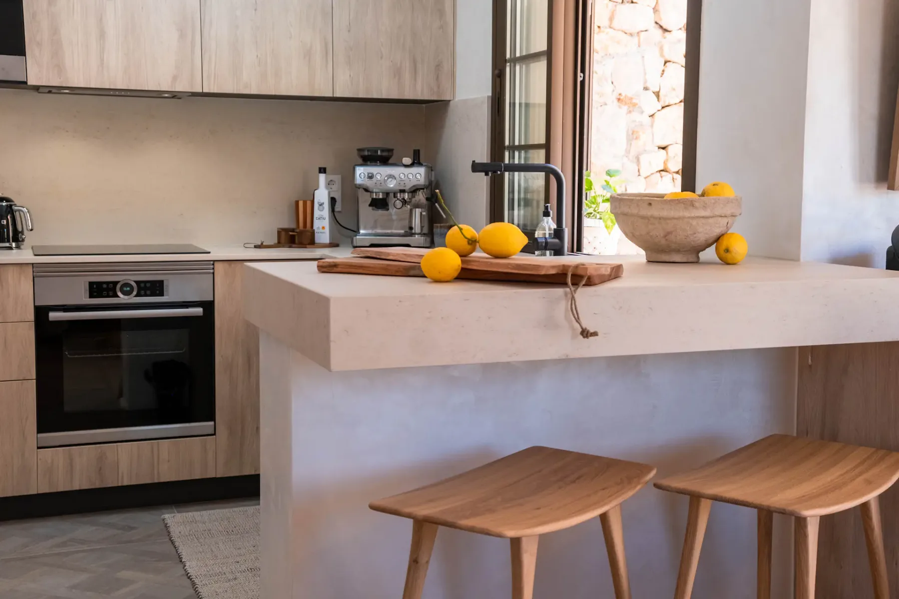 A modern kitchen with a wide counter, wooden stools, and citrus fruit.