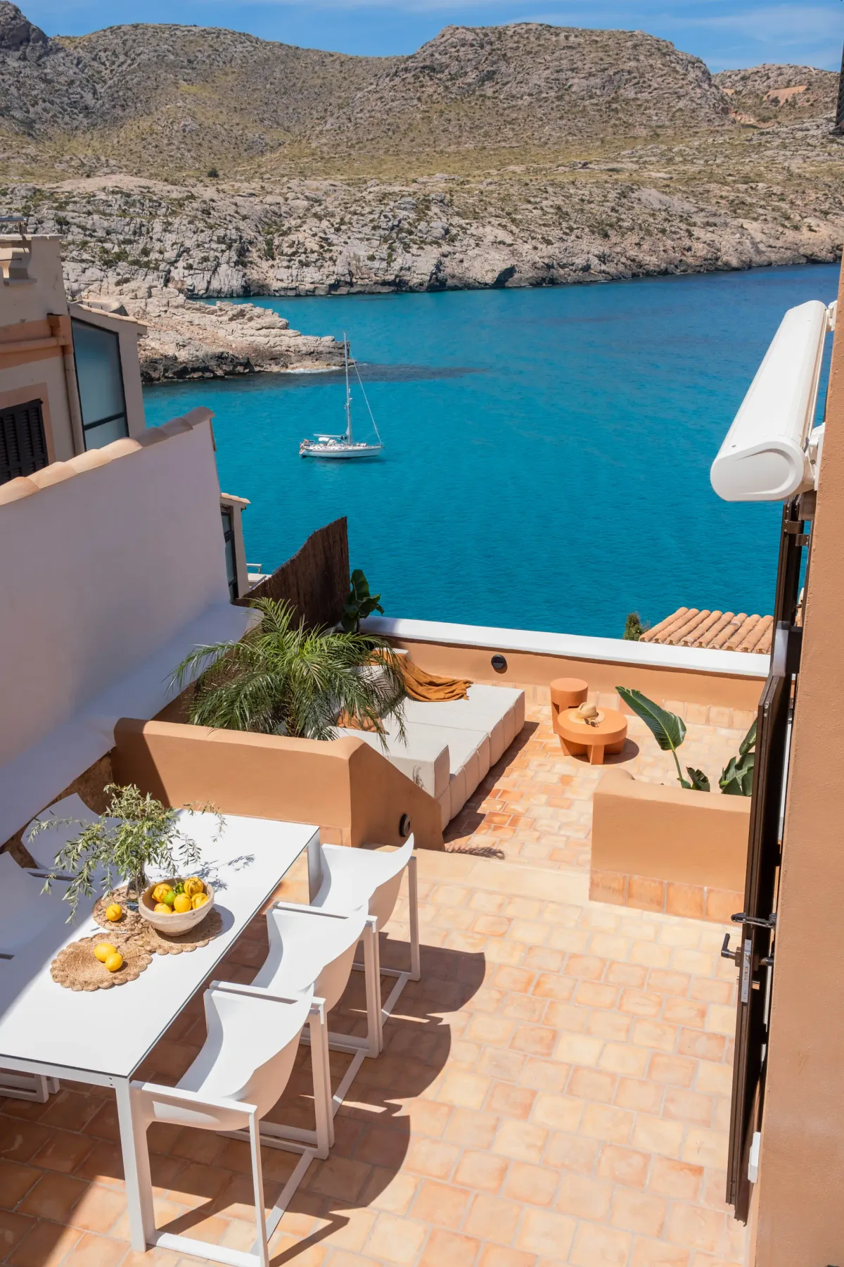 A tiled outdoor terrace overlooks turquoise water, a rocky coastline, and a sailboat.