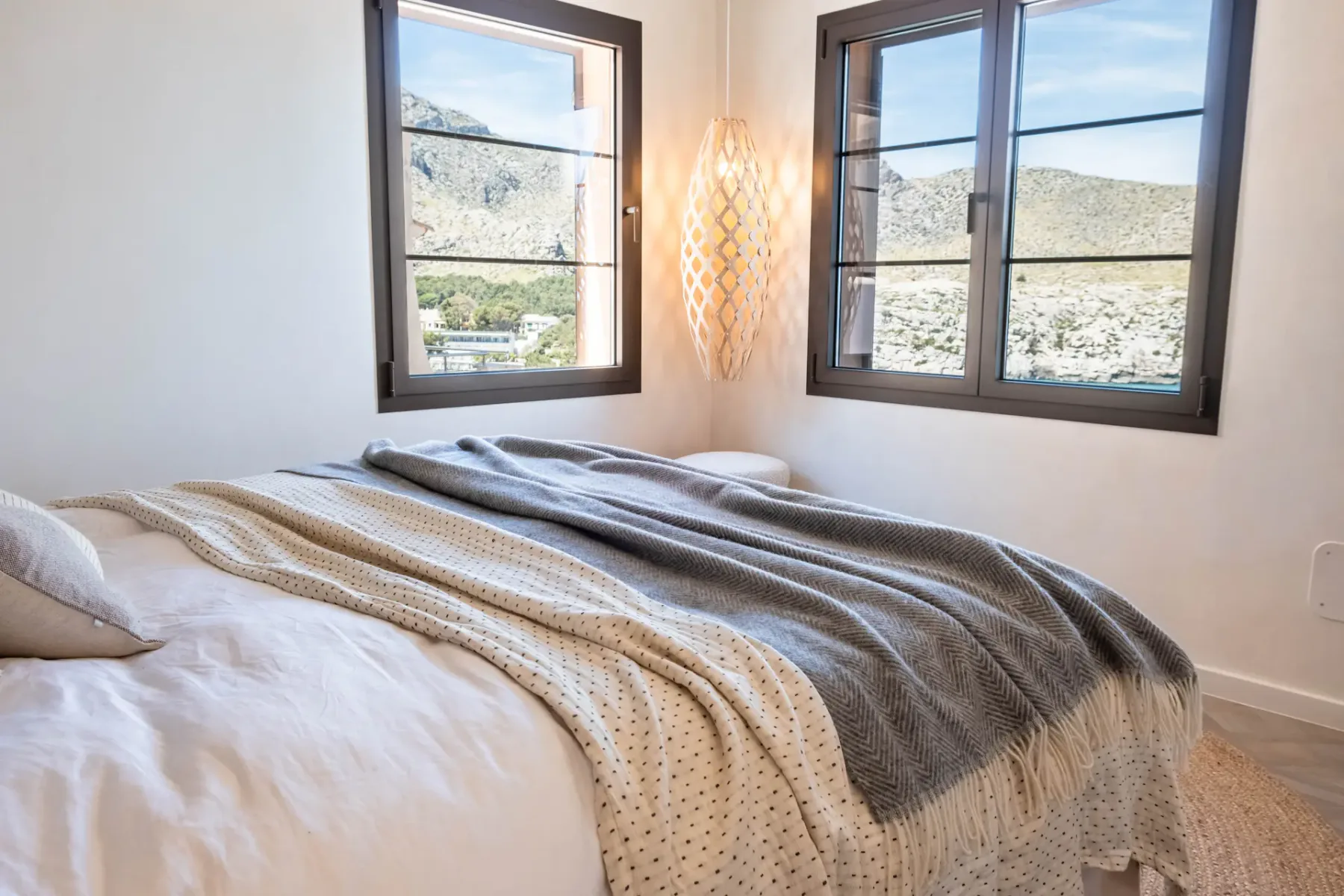 A bedroom with layered bedding and windows overlooking a hillside.
