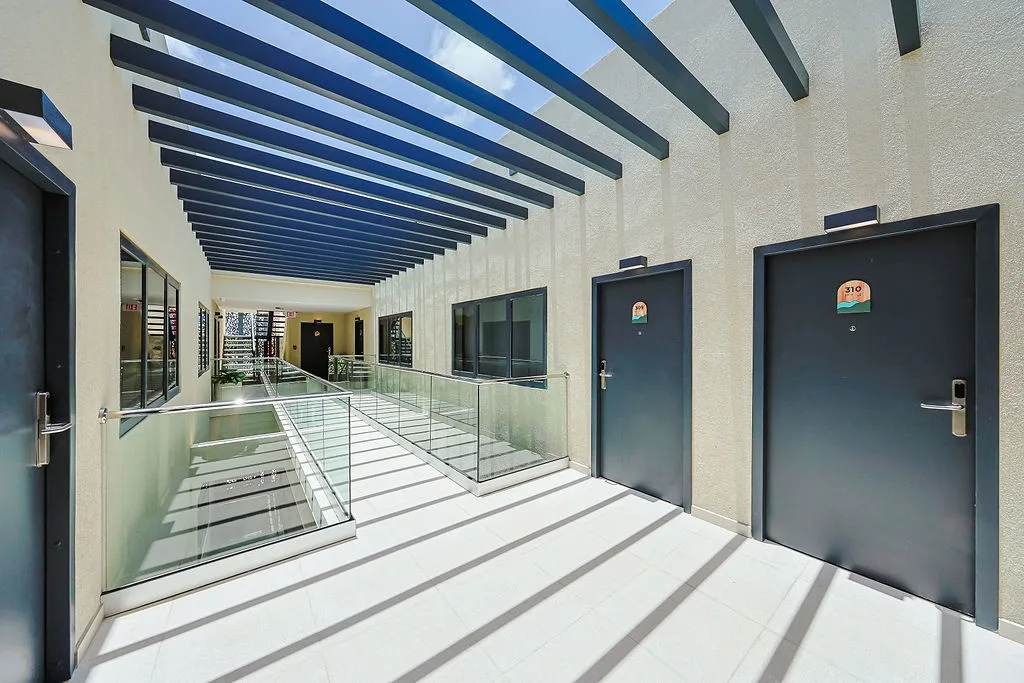 The open-air hallway showcases modern design with striking light and shadow patterns cast through the slatted pergola overhead. Clean lines and glass railings create a bright, airy passage connecting your private space to the rest of the property.