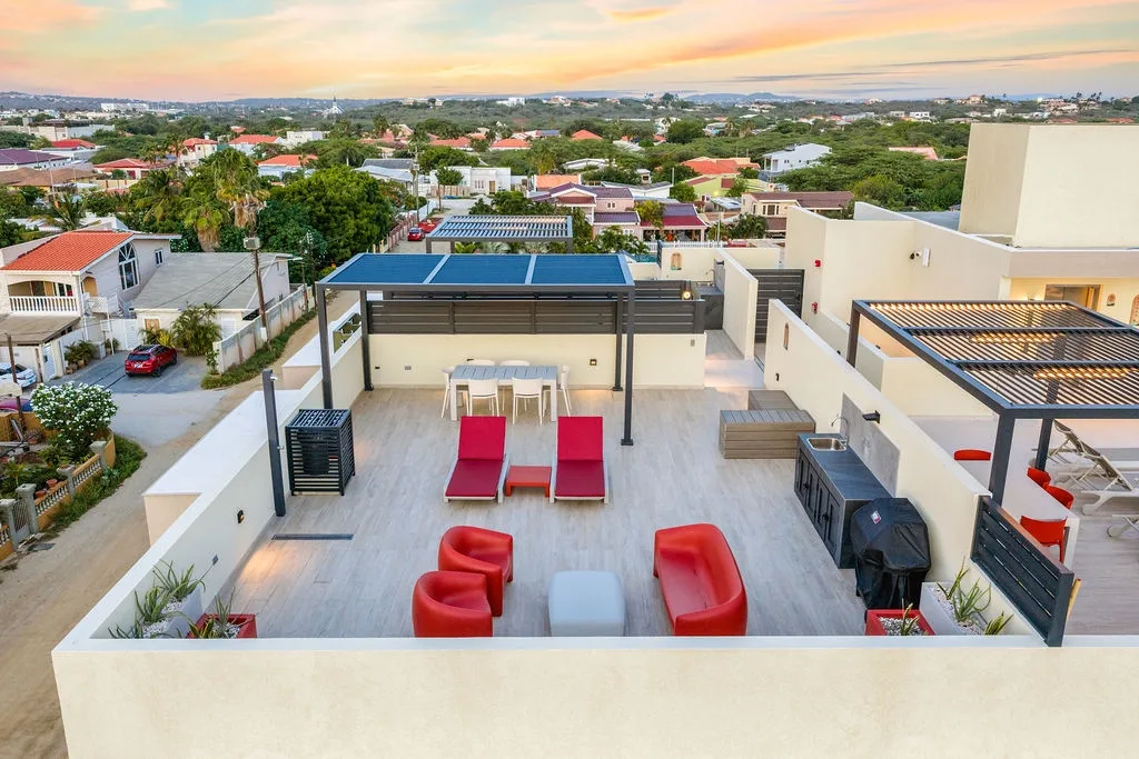 Rooftop terrace with lounge seating and dining area overlooking a neighborhood of red-roofed homes and lush greenery at sunset.