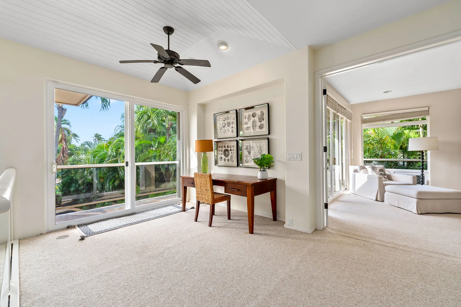 A bright carpeted room has a desk by large windows and an open doorway to a sitting area.