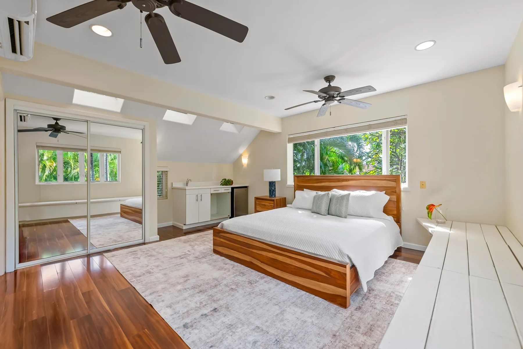 A bright bedroom with a large bed, wood headboard, and windows overlooking greenery.