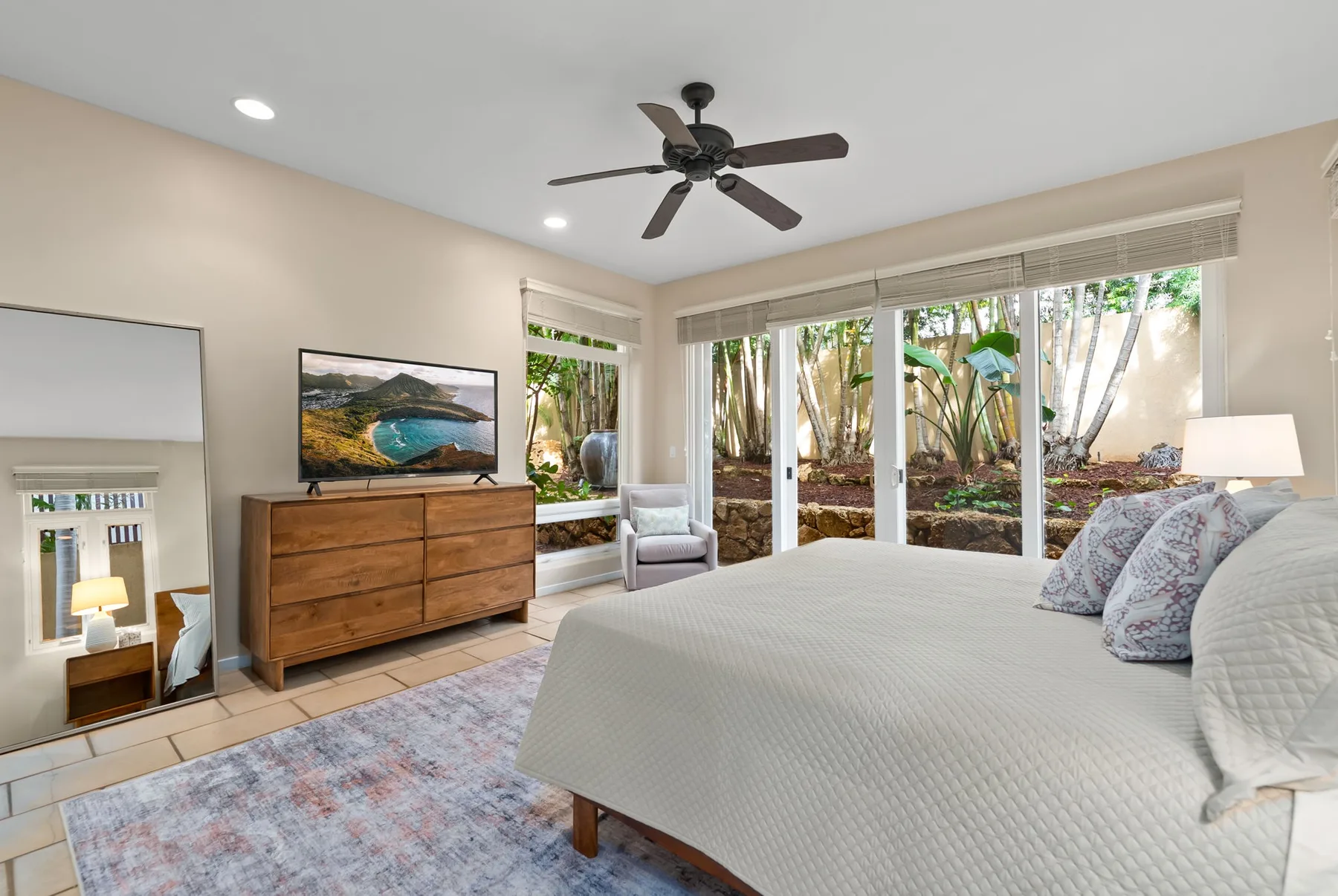 A bright bedroom has a large bed facing a dresser and television beside wide windows.