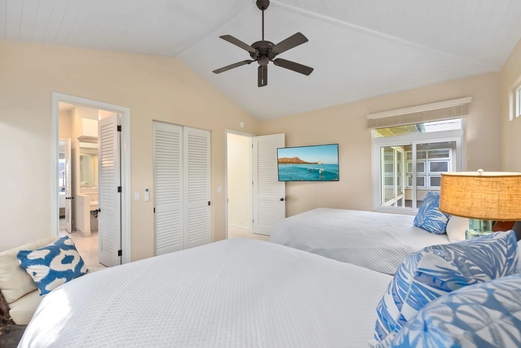 A bright bedroom with two beds, blue patterned pillows, and a wall-mounted television.