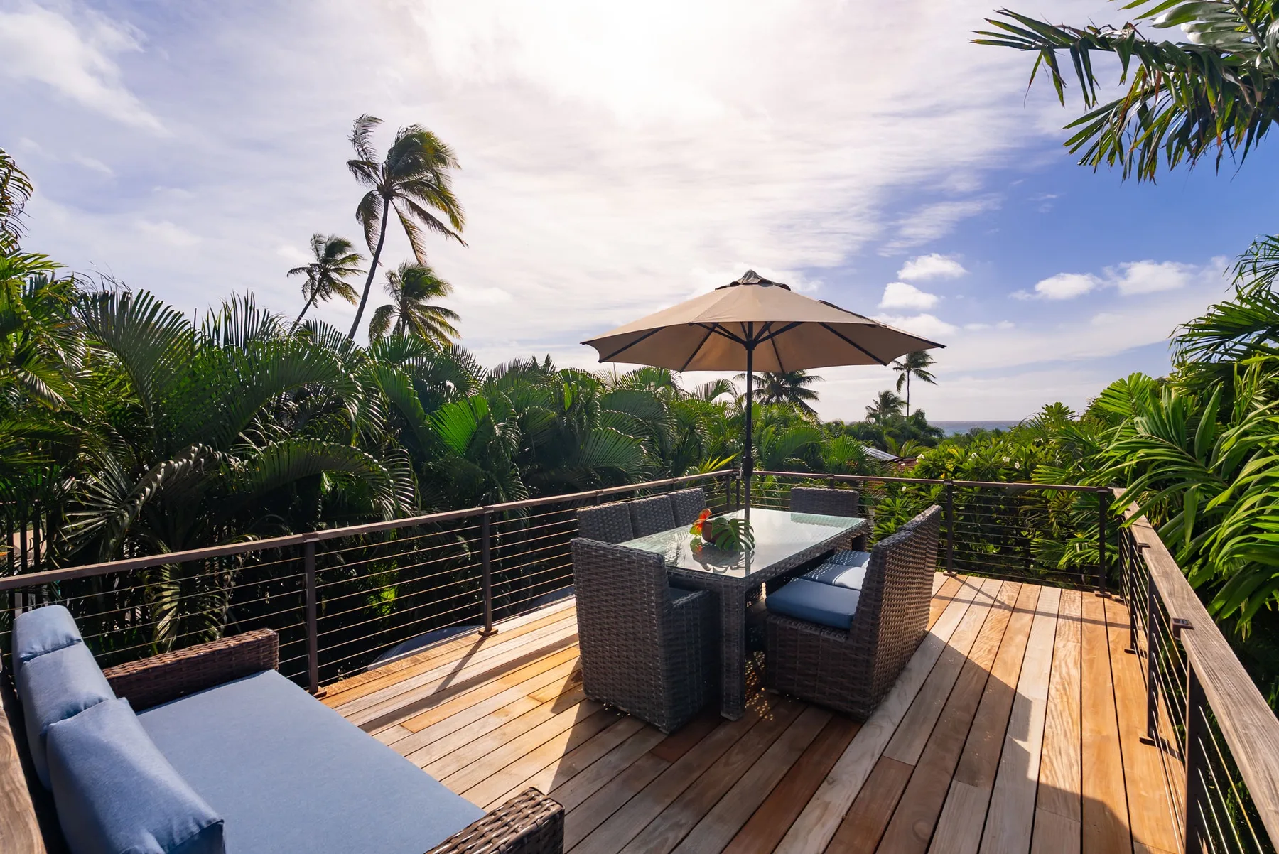 A tropical outdoor deck with a dining table, umbrella, and surrounding greenery.
