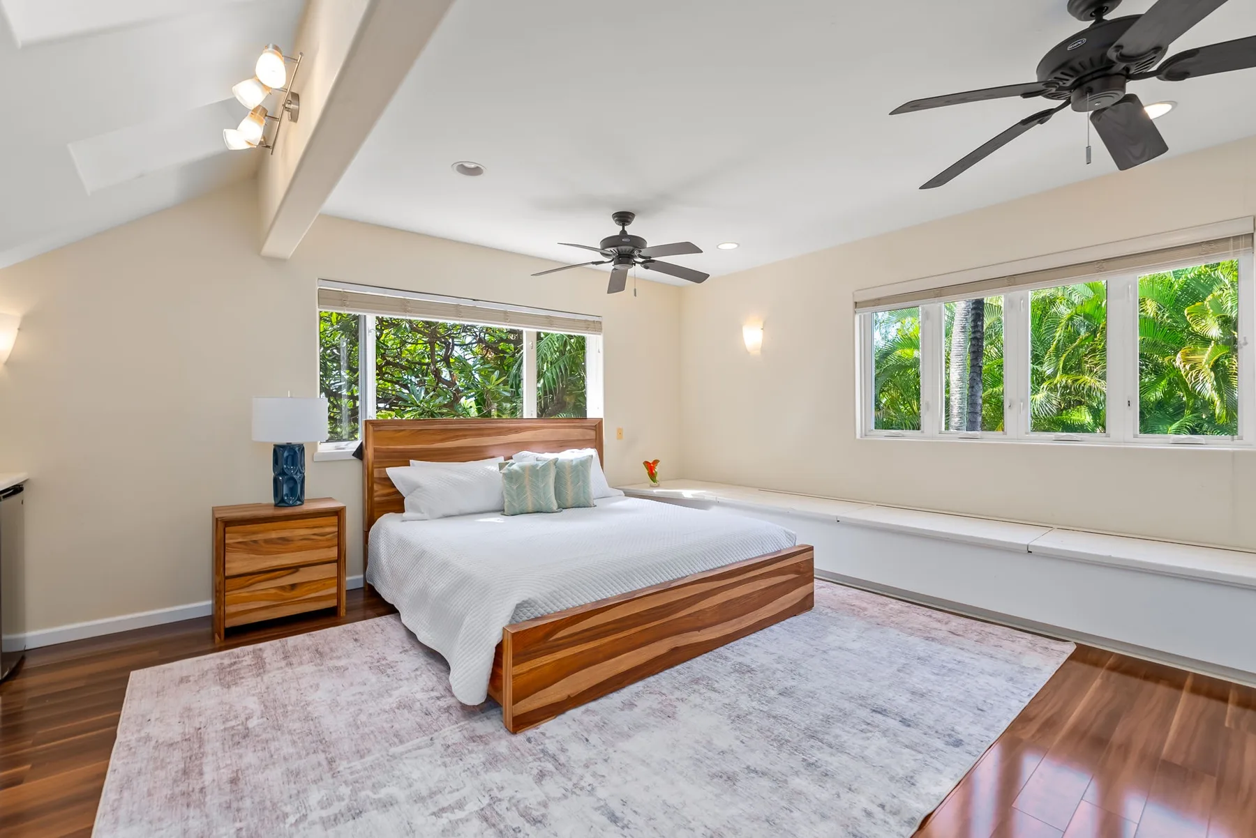 A bright bedroom with a wooden bed, pale rug, and windows overlooking greenery.