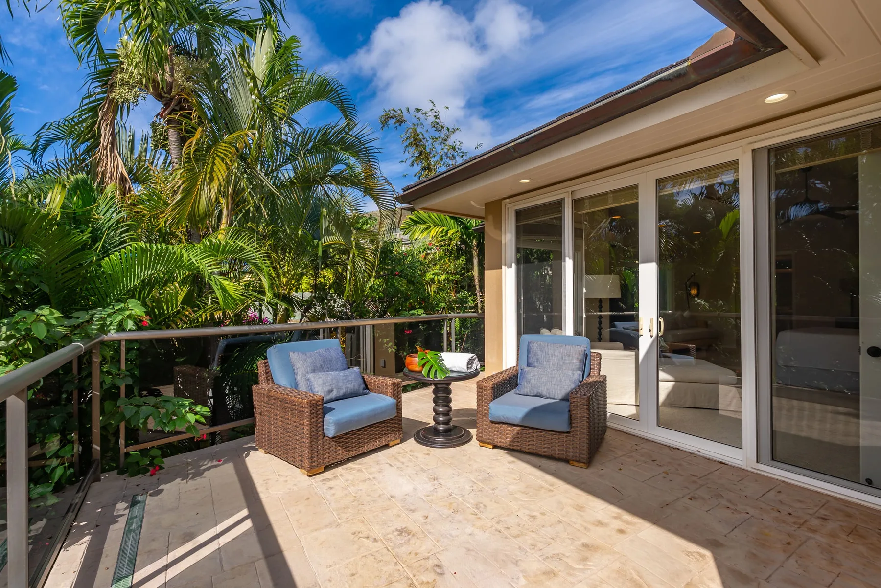 A sunny patio with two cushioned seats beside large glass doors and tropical greenery.