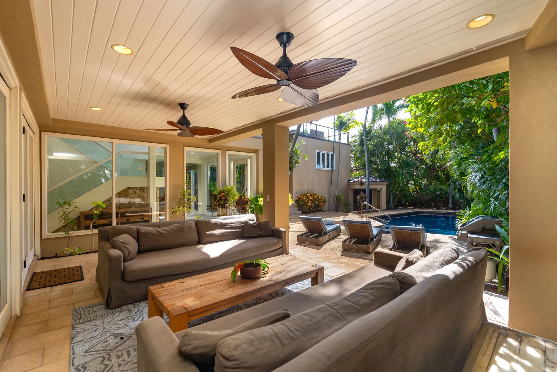 A covered patio lounge with seating around a wooden coffee table, overlooking a pool.