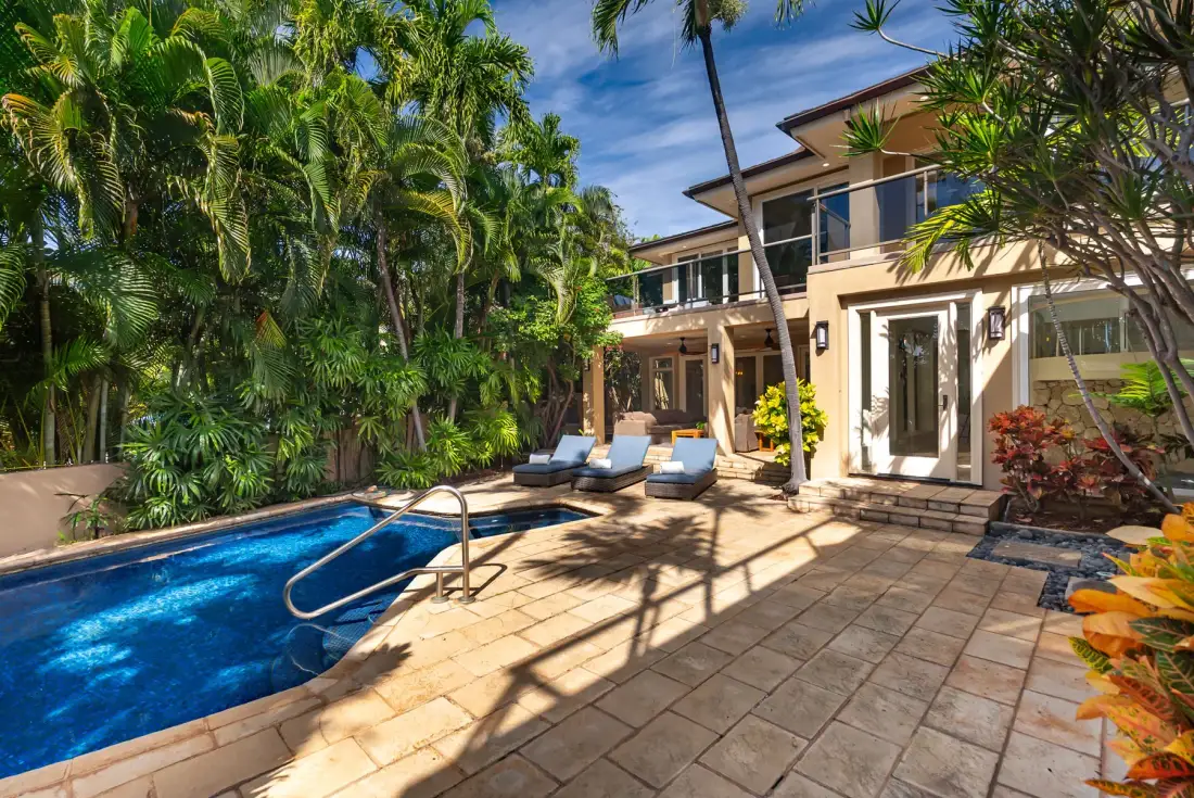 A swimming pool and paved patio sit beside a two-story house surrounded by tropical plants.
