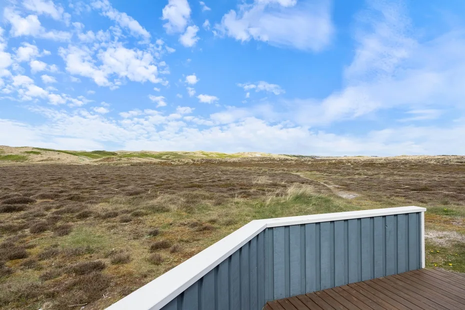 Una terraza de madera da a un paisaje amplio y bajo bajo un cielo azul con nubes.
