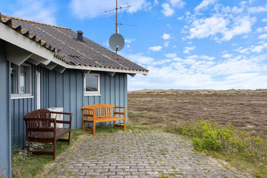 Una casa pequeña con asientos al aire libre junto a un camino pavimentado y un paisaje abierto.